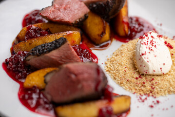 Slices of roasted duck breast lying on caramelized apple slices, covered in berry sauce and served with bread crumbs and a dollop of cream cheese, creating a gourmet culinary experience