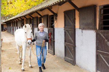 Mature woman jockey leading her white corse out of stable for ride.