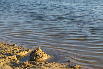 sandy beach on a calm body of water, a lake. a small sandcastle, built right on the shoreline. The water is calm, and its surface gently ripples, reflecting the sunlight.