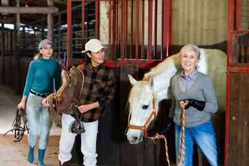 Positive Asian horsewoman holding saddle while elderly female stable keeper leading white horse out...
