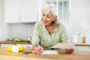 Smiling elderly woman standing near countertop at kitchen at home, reading papers