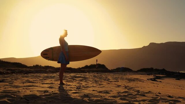 LENS FLARE, SILHOUETTE: Young woman stands on beach at sunset holding a wooden surfboard. She is checking the waves at a surf spot before heading into water for an evening session on Canary Islands.