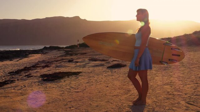 LENS FLARE, PORTRAIT: Young woman is restlessly standing on shore watching waves. With a wooden surfboard in hands, she hesitates slightly before heading out for a morning surf session on Lanzarote.
