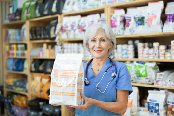 Experienced veterinarian senior woman is standing in pet store with pack of food in hands. Specialist offers services selection of medical and dietary nutrition for pet