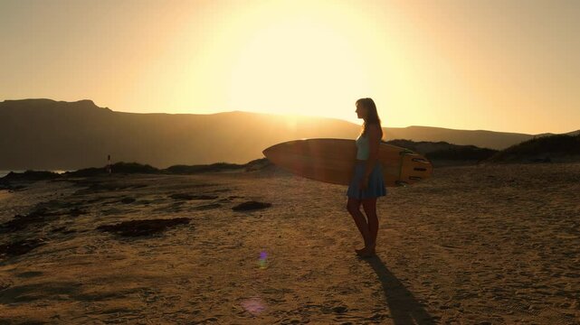 LENS FLARE, SILHOUETTE: Female surfer stands on beach watching beautiful waves breaking in offshore winds. Woman in a skort and with a surfboard in hands is getting ready for a morning surf session.