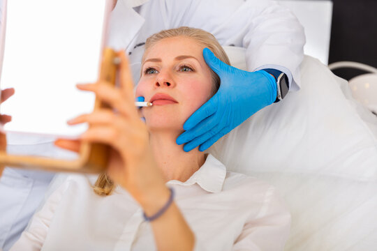 Cosmetologist planning hardware treatments to young female client, applying marks with white pencil on her face while woman looking in mirror