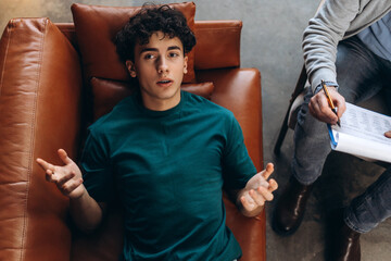 Top view of psychologist holding clipboard in his hands, teenage boy lying on couch