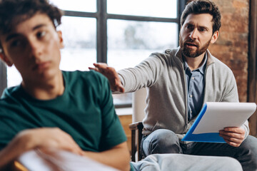 Male psychotherapist talking to troubled teenage boy sitting on couch
