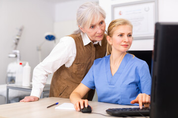 Obraz premium Young woman dermatologist using computer during appointment. Her patient, senior woman, standing near and looking at display.