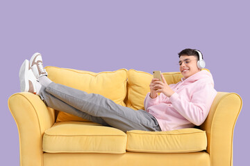 Young man in headphones using mobile phone on sofa against lilac background