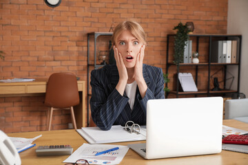 Young shocked female accountant sitting at workplace with laptop and documents in office