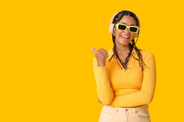 Young African-American woman listening to music and pointing at something on yellow background