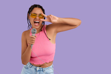Young African-American woman in sunglasses with microphone singing on lilac background