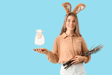 Beautiful young woman in bunny ears with pussy willow branches and toy sheep on blue background