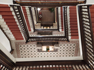 Red carpeted staircases in a hotel form a geometric pattern as they spiral downward. Wooden railings with elaborate designs frame the stairs, creating a repetitive motif.