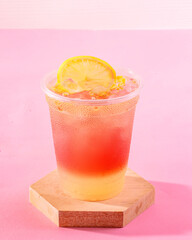 A glass of pink lemonade with a slice of lemon on top. The drink is served in a plastic cup and is sitting on a wooden coaster