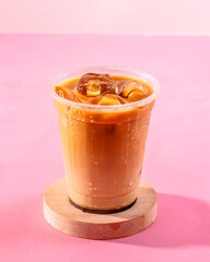 A cup of coffee with ice cubes in it sits on a wooden coaster. The coaster is on a pink background