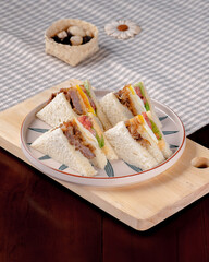 A plate of four sandwiches sits on a wooden board. The sandwiches are cut in half and are filled with various ingredients, including meat, cheese, and vegetables. The plate is placed on a table