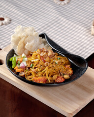 A bowl of food with noodles and vegetables on a wooden table. The bowl is black and the table is brown