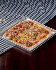 A square pizza with a variety of toppings including mushrooms, corn, and peppers. The pizza is sitting on a table with a checkered tablecloth