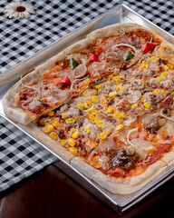 A pizza with corn and peppers on it is sitting on a table. The pizza is cut into slices and is placed in a metal pan