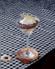 A dessert in a glass with a leaf on top. The dessert is on a wooden coaster. The dessert is on a checkered tablecloth