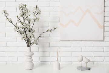 Vase of white blooming branches with decor on dresser near white brick wall in living room