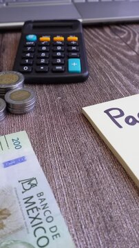 Toma vertical de escritorio con nota adhesiva con la palabra pagos, con billete de 200 pesos mexicanos y monedas apilada, al fondo calculadora