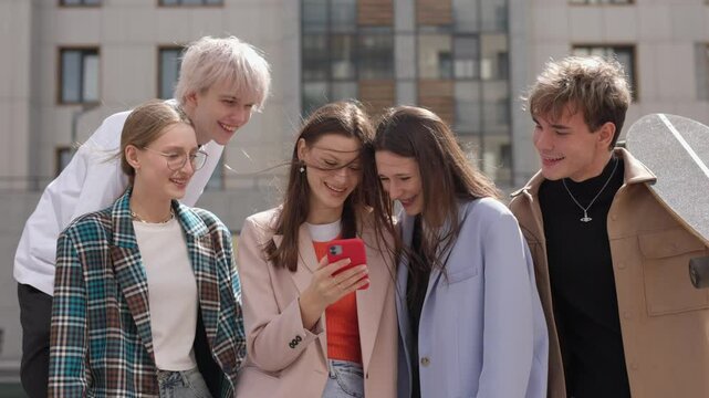 A group of international young friends laughs together, gathered in a circle as they scroll through their phones. With smiles on their faces, they watch funny videos online, caught up in the