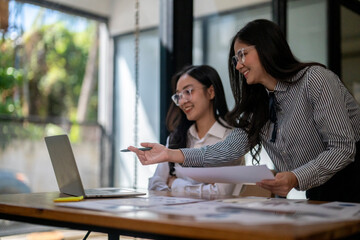 Fototapeta premium Two women are sitting at a desk with a laptop and a piece of paper