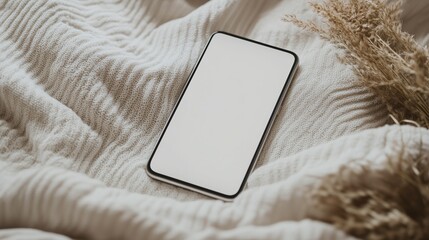 Smartphone with a blank screen resting on a soft knitted blanket with dried flowers. Cozy digital lifestyle concept.