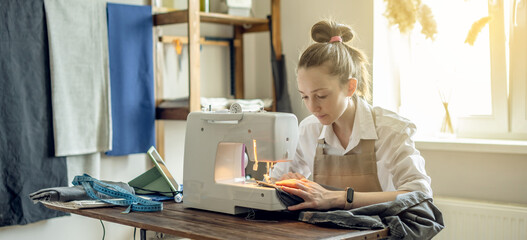 A female tailor is working on a sewing machine stitching fabric in sunny room. Home sewing...