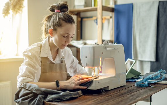 A female tailor is working on a sewing machine stitching fabric in sunny room. Home sewing production