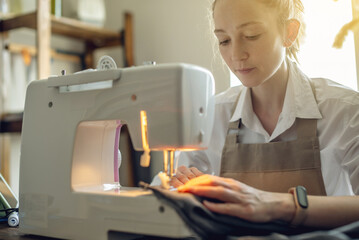 A female tailor is working on a sewing machine stitching fabric in sunny room. Home sewing production
