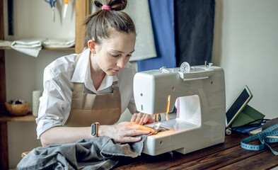 A female tailor is working on a sewing machine stitching fabric in sunny room. Home sewing...