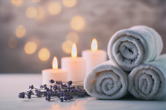 The image shows a serene spa setting with rolled white towels, lit candles, and a sprig of lavender, all against a softly focused background with warm lights