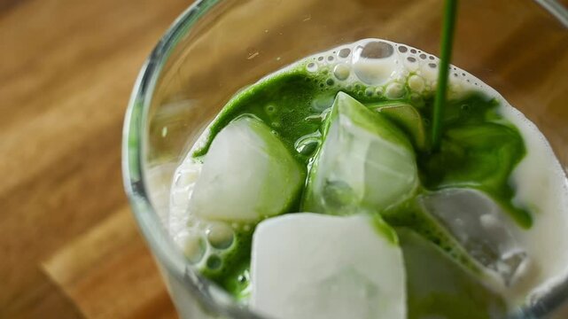 Slow motion video of organic green Japanese matcha being poured into a glass filled with ice cubes and fresh milk creating a refreshing summer iced matcha latte indoors with soft natural light.