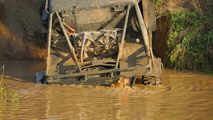 Close-up of the rear part of an SUV stuck in a muddy ditch. The vehicle is partially submerged in mud, the cooling system and the safety cage are visible.