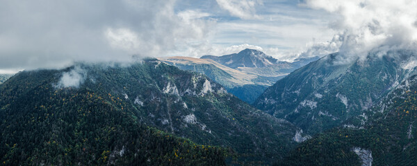 A breathtaking panorama of a mountainous landscape shrouded in mist. Lush greenery slopes into misty valleys beneath a dynamic sky, creating a serene yet dramatic view. Panorama of mountain range.