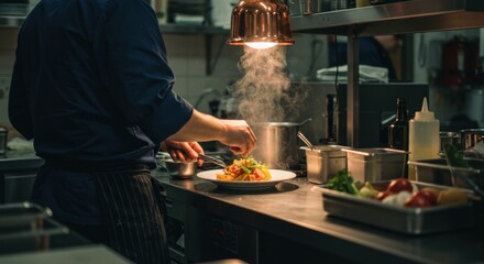 A chef is skillfully plating a colorful gourmet dish under the warm glow of pendant lighting in a busy kitchen. The space is filled with stainless steel counters and various ingredients carefully
