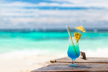 The image features a vibrant blue cocktail garnished with a pineapple wedge and a small umbrella, placed on a wooden table against a blurred beach backdrop with clear turquoise waters and a blue sky