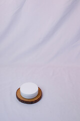 A minimalist still life captures a small, round, white cosmetic jar resting on a natural, circular wood.