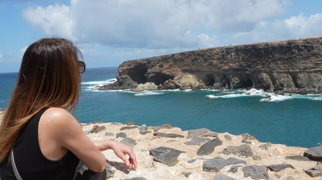 A woman with long dark hair admires the Ajui Caves on the island of Fuerteventura from the shore. Canary Islands, Spain.