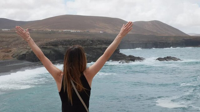 A woman with long, dark hair raises her arms as a symbol of freedom and joy on the Atlantic Ocean coast. Fuerteventura Island, Ajui Caves. Canary Islands, Spain.