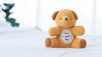 Little teddy bear toy is cute, sitting alone on a comfortable white bed, white pillow and white duvet cover in bedroom at home in morning.