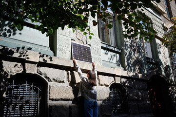 Woman holding photovoltaic solar panel in front of historical building, wearing plaid shirt and...