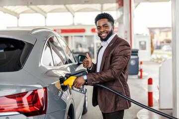 Portrait of happy interracial man standing at gas station and filling up tank with gasoline.