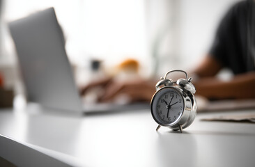 close up photograph of silver alarm clock desk, with blurred background of person working laptop. scene conveys sense