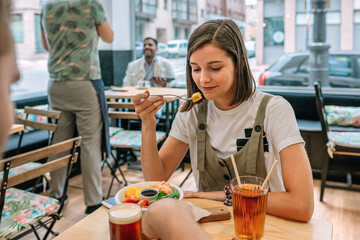 Portrait of smiling young woman tasting a piece of mango from her poke bowl while having lunch with a friend in a modern restaurant, enjoying a healthy and delicious meal together