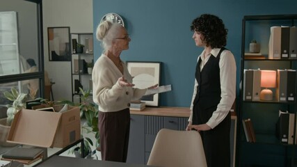 Medium shot of elderly woman briefing younger colleague taking her place, handing over final documents and sharing last pieces of advice before officially retiring from her role in office